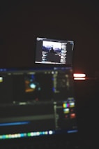A dimly lit room with two computer screens displaying editing software. The foreground monitor shows various color and timeline adjustments, while the background monitor displays a video of a person in front of a large landscape. The ambiance is focused and technology-oriented.