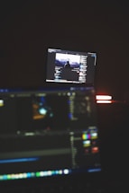 A dimly lit room with two computer screens displaying editing software. The foreground monitor shows various color and timeline adjustments, while the background monitor displays a video of a person in front of a large landscape. The ambiance is focused and technology-oriented.