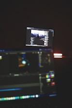 A dimly lit room with two computer screens displaying editing software. The foreground monitor shows various color and timeline adjustments, while the background monitor displays a video of a person in front of a large landscape. The ambiance is focused and technology-oriented.