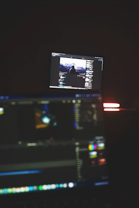 A dimly lit room with two computer screens displaying editing software. The foreground monitor shows various color and timeline adjustments, while the background monitor displays a video of a person in front of a large landscape. The ambiance is focused and technology-oriented.