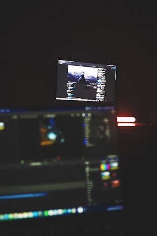 A dimly lit room with two computer screens displaying editing software. The foreground monitor shows various color and timeline adjustments, while the background monitor displays a video of a person in front of a large landscape. The ambiance is focused and technology-oriented.