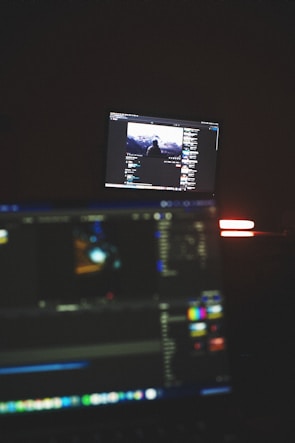 A dimly lit room with two computer screens displaying editing software. The foreground monitor shows various color and timeline adjustments, while the background monitor displays a video of a person in front of a large landscape. The ambiance is focused and technology-oriented.
