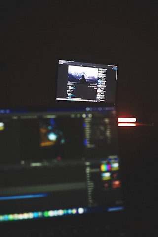 A dimly lit room with two computer screens displaying editing software. The foreground monitor shows various color and timeline adjustments, while the background monitor displays a video of a person in front of a large landscape. The ambiance is focused and technology-oriented.