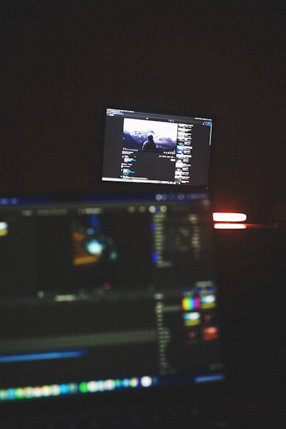 A focused video editor working intently at a dual-monitor setup with editing software open, bathed in warm orange light.