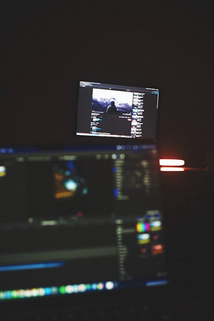 A dimly lit room with two computer screens displaying editing software. The foreground monitor shows various color and timeline adjustments, while the background monitor displays a video of a person in front of a large landscape. The ambiance is focused and technology-oriented.