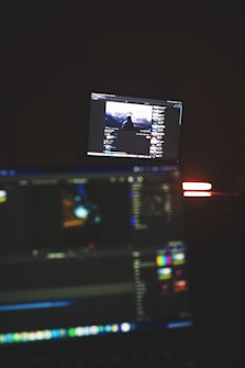 A dimly lit room with two computer screens displaying editing software. The foreground monitor shows various color and timeline adjustments, while the background monitor displays a video of a person in front of a large landscape. The ambiance is focused and technology-oriented.