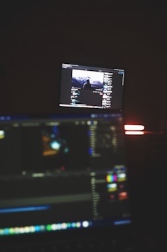 A dimly lit room with two computer screens displaying editing software. The foreground monitor shows various color and timeline adjustments, while the background monitor displays a video of a person in front of a large landscape. The ambiance is focused and technology-oriented.