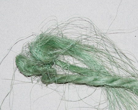 A close-up image of a bundle of fibrous material, with twisted and loose strands. The fibers appear green and have a shiny, textured appearance against a light background.