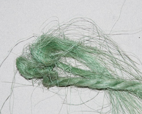 A close-up image of a bundle of fibrous material, with twisted and loose strands. The fibers appear green and have a shiny, textured appearance against a light background.