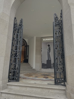 An elegant stone building entrance featuring ornate black wrought iron gates, marble steps, and a polished mosaic floor. Inside, a large Burberry advertisement displays a fashion model. The space is well-lit with recessed ceiling lights.
