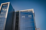A modern office building with a glass facade reflecting the clear blue sky. The structure is composed of multiple stories with large, rectangular panes of glass and grey metal accents. The architectural design features sharp lines and a sleek, contemporary aesthetic.