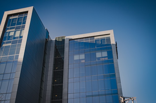 A modern office building with a glass facade reflecting the clear blue sky. The structure is composed of multiple stories with large, rectangular panes of glass and grey metal accents. The architectural design features sharp lines and a sleek, contemporary aesthetic.