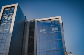 A modern office building with a glass facade reflecting the clear blue sky. The structure is composed of multiple stories with large, rectangular panes of glass and grey metal accents. The architectural design features sharp lines and a sleek, contemporary aesthetic.