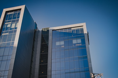 A modern office building with a glass facade reflecting the clear blue sky. The structure is composed of multiple stories with large, rectangular panes of glass and grey metal accents. The architectural design features sharp lines and a sleek, contemporary aesthetic.