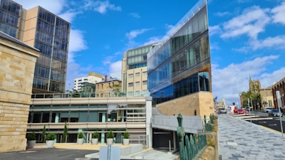 A modern architectural scene with a prominent glass building reflecting the blue sky and adjacent structures. The area is urban, with multiple buildings featuring different architectural styles. The path beside the buildings is paved, leading towards a street with visible traffic lights and road signs.