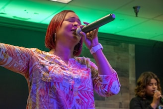 A woman with short red hair is passionately singing into a microphone. She is wearing a colorful patterned blouse and a white watch on her left wrist. Her left arm is raised as she closes her eyes while singing. In the background, another woman is also holding a microphone, appearing to sing or speak.