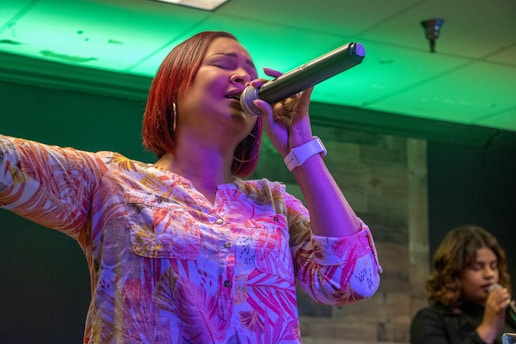 A woman with short red hair is passionately singing into a microphone. She is wearing a colorful patterned blouse and a white watch on her left wrist. Her left arm is raised as she closes her eyes while singing. In the background, another woman is also holding a microphone, appearing to sing or speak.