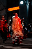 A woman in vibrant traditional attire dances with a broad smile. Her outfit includes a glowing shawl adorned with lights, a polka dot dress with vivid red ruffles, and she is moving energetically. The background shows another person dressed similarly, with blurred street lights and signs creating a festive atmosphere.