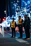 A nighttime street scene with several people standing, two of whom are wearing high-visibility vests marked 'CADET'. The background features illuminated trees decorated with blue lights, creating a festive atmosphere.