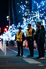 A nighttime street scene with several people standing, two of whom are wearing high-visibility vests marked 'CADET'. The background features illuminated trees decorated with blue lights, creating a festive atmosphere.