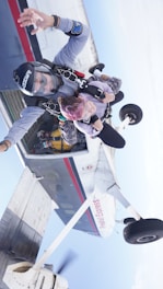 A tandem skydive is taking place as two individuals exit an airplane in mid-air. One person is attached to a professional with parachute gear, as they freefall with the sky visible in the background. The airplane has visible propellers and a distinct white, red, and blue color scheme.