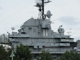 A large, gray naval battleship is pictured, featuring radar and communication equipment on its upper structure. The ship's superstructure includes multiple antennas, satellite dishes, and various electronic devices. In the foreground, green trees partially obscure the lower part of the ship.