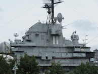A large, gray naval battleship is pictured, featuring radar and communication equipment on its upper structure. The ship's superstructure includes multiple antennas, satellite dishes, and various electronic devices. In the foreground, green trees partially obscure the lower part of the ship.