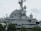 A large, gray naval battleship is pictured, featuring radar and communication equipment on its upper structure. The ship's superstructure includes multiple antennas, satellite dishes, and various electronic devices. In the foreground, green trees partially obscure the lower part of the ship.