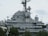 A large, gray naval battleship is pictured, featuring radar and communication equipment on its upper structure. The ship's superstructure includes multiple antennas, satellite dishes, and various electronic devices. In the foreground, green trees partially obscure the lower part of the ship.