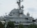 A large, gray naval battleship is pictured, featuring radar and communication equipment on its upper structure. The ship's superstructure includes multiple antennas, satellite dishes, and various electronic devices. In the foreground, green trees partially obscure the lower part of the ship.