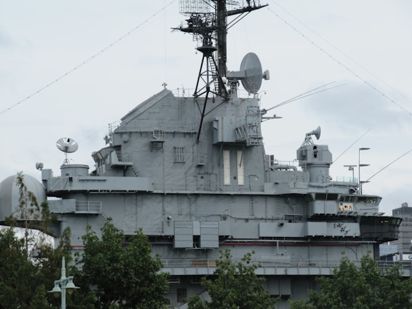 A large, gray naval battleship is pictured, featuring radar and communication equipment on its upper structure. The ship's superstructure includes multiple antennas, satellite dishes, and various electronic devices. In the foreground, green trees partially obscure the lower part of the ship.