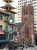 A bustling urban street scene featuring a blend of architectural styles, with a prominent red brick church tower exhibiting gothic elements alongside a modern beige and blue building with Asian architectural influences. The scene includes pedestrians, traffic lights, and signage welcoming visitors to Chinatown.