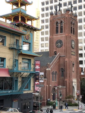A bustling urban street scene featuring a blend of architectural styles, with a prominent red brick church tower exhibiting gothic elements alongside a modern beige and blue building with Asian architectural influences. The scene includes pedestrians, traffic lights, and signage welcoming visitors to Chinatown.