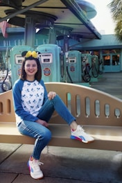A smiling person sitting on a bench in front of vintage-style gas pumps, wearing a hat with mouse ears and a bow, a white and blue Mickey Mouse shirt, jeans, and colorful sneakers.