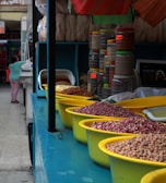 A market stall featuring a variety of legumes and grains displayed in yellow bowls on a blue counter. Behind the counter are stacked containers filled with different ingredients. A person, partially visible, appears to be interacting with another stall in the background.