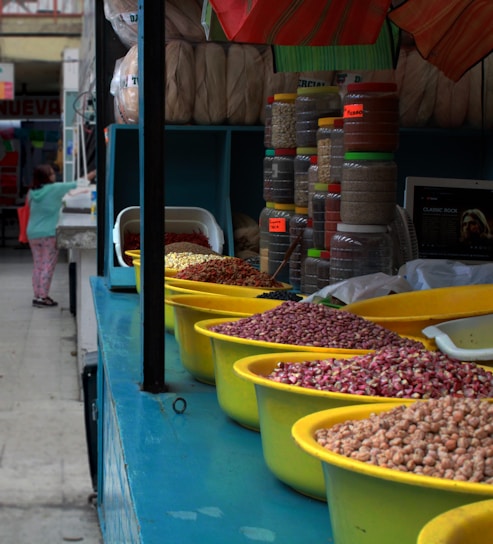 A market stall featuring a variety of legumes and grains displayed in yellow bowls on a blue counter. Behind the counter are stacked containers filled with different ingredients. A person, partially visible, appears to be interacting with another stall in the background.