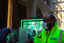 A person wearing a bright neon yellow jacket and a face mask stands in the foreground inside a modern building with large glass windows and dark walls. Behind them, a digital display screen shows information related to 'Green energy for London.'. People are seen using exercise bikes, partially visible to the left of the screen.