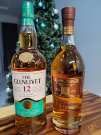 Two bottles of single malt Scotch whisky are placed on a wooden surface. The bottle on the left is The Glenlivet 12 years, featuring a teal and white label with a gold coin detail. The bottle on the right is Glenmorangie 18 years, with a dark brown label and gold accents. Behind them, a decorated Christmas tree can be seen, adorned with blue and white ornaments.