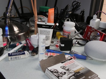 A cluttered desk with various items including skincare products, a jar of pills, a box labeled Nyquil, a red digital camera, a small white speaker, pens, cables, and a lamp. There's also a pack of cigarettes in the foreground.