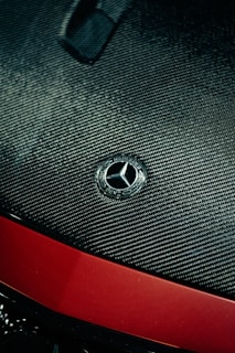 A close-up view of a car hood featuring a carbon fiber texture. The Mercedes-Benz emblem is prominently displayed in silver at the center. Below the emblem, the hood transitions to a glossy red finish.