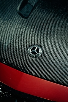 A close-up view of a car hood featuring a carbon fiber texture. The Mercedes-Benz emblem is prominently displayed in silver at the center. Below the emblem, the hood transitions to a glossy red finish.