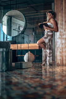 A woman stands leaning against a wooden partition in a modern, stylish interior. She is absorbed in reading a book, dressed in a relaxed, fashionable two-piece outfit. The space around her features a round mirror, a ceramic vase, and polished patterned flooring that reflects light softly. The overall ambiance of the room is contemporary and serene with a touch of elegance.