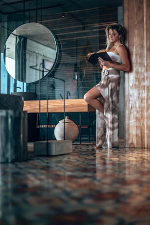A woman stands leaning against a wooden partition in a modern, stylish interior. She is absorbed in reading a book, dressed in a relaxed, fashionable two-piece outfit. The space around her features a round mirror, a ceramic vase, and polished patterned flooring that reflects light softly. The overall ambiance of the room is contemporary and serene with a touch of elegance.