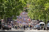 A large crowd of people is gathered in the middle of a street lined with green trees, participating in a protest or demonstration. Numerous signs and banners with different messages are held up. Some flags are visible among the crowd. Cars are parked alongside the street, and the scene suggests a lively and energetic atmosphere.