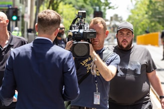 A cameraman is filming an interview on a city street, focused on a man in a suit who is facing away from the camera. Another man stands nearby, looking towards the camera. The scene is lively, with green trees and a yellow barricade visible in the background.
