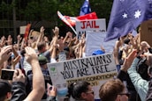 A large crowd of people with various signs and banners are gathered together, raising their hands in the air. One prominent sign reads 'Australians you are free...' with a drawing that includes a face in a mask. Flags with stars and writing are visible among the crowd, and there is greenery in the background.