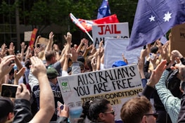 A large crowd of people with various signs and banners are gathered together, raising their hands in the air. One prominent sign reads 'Australians you are free...' with a drawing that includes a face in a mask. Flags with stars and writing are visible among the crowd, and there is greenery in the background.