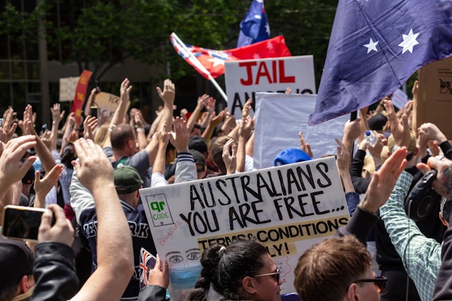 A large crowd of people with various signs and banners are gathered together, raising their hands in the air. One prominent sign reads 'Australians you are free...' with a drawing that includes a face in a mask. Flags with stars and writing are visible among the crowd, and there is greenery in the background.