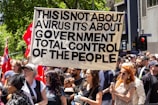 A crowd of people are participating in a protest, holding a large sign that reads 'This is not about a virus its about government total control of the people.' The protestors are diverse and appear engaged. There are red flags with stars in the background, and the setting is urban with trees and buildings visible.