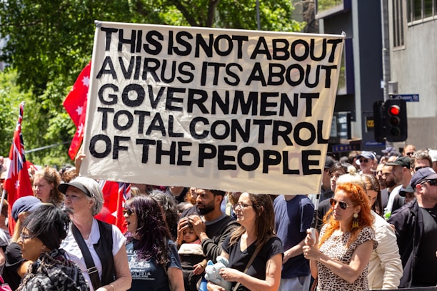 A crowd of people are participating in a protest, holding a large sign that reads 'This is not about a virus its about government total control of the people.' The protestors are diverse and appear engaged. There are red flags with stars in the background, and the setting is urban with trees and buildings visible.